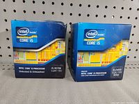 Front view of two Intel Core i5 processors boxes (i5-3570K and i5-2405S) on gray surface with pegboard background.