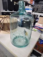 Full view of the 5 gallon glass jug showing clear to aqua tint, round body, and embossed text on the front.