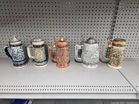 Front view of five Avon beer steins in a row with distinct designs and metal lids on a white shelf with pegboard background.