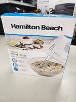 Front view of box showing hand mixer mixing bowl of cookie dough and desserts in the background