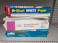 Three packages of printer paper stacked on shelf including Canon Brilliant White Paper, IBM Premium Document Ink Jet Paper, and Double Side Photo Paper