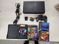 Full lot overview showing the PS2 console, controller, power adapter, AV cables, two memory cards, and four game discs with cases scattered around the console.