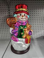 Front view of Thomas Pacconi snowman figurine showing red hat, scarf, wooden 'Let It Snow' sign and gingerbread man cookie in white snow body on round wooden base.