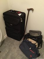 Black Air Canada carry-on roller bag standing upright with two additional smaller bags nearby and a wooden cane leaning against the wall