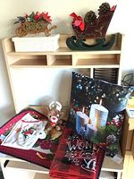 Photo showing Christmas pillow with candles, small reindeer figurine, wooden sled decoration with pine cones on top of a hutch shelf, and a fabric piece with Christmas embroidery.