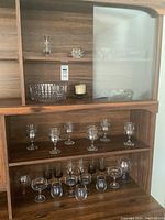 Full view of wood cabinet shelf showcasing multiple crystal and glass stemware and serveware items including wine glasses, brandy snifters, bowls, and candle.