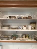 Full shelf with assorted dishware including stacked plates, bowls, various mugs, and two crystal vases.