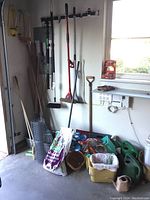 Photo showing corner wall with hanging garden tools including shovels, brooms, and hedge trimmers. Below on floor are watering cans, soil bag, gloves, bins with small tools, and hose attachment.