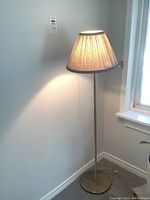 Brass floor lamp with beige conical shade standing near a wall and window.