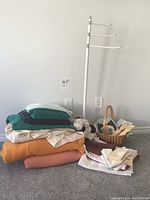 Photo showing stacked various linens including towels, wool blankets, a wicker basket with linens inside, a small sheep plush, and a white metal floor-standing towel rack with four arms.
