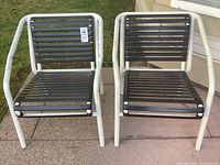 Two outdoor chairs side by side featuring white metal tubular frames and dark slatted seats and backs on a concrete surface with some wear visible on frames.