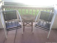 Two white metal framed outdoor chairs with black plastic slat seats and backs. Signs of wear are visible on the metal frames. Between them is a round wicker table.