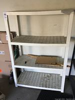 Front view of white plastic four shelf storage rack in storage area, with cardboard and small items on middle shelf.