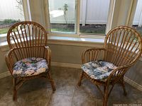 Pair of rattan chairs placed near a window with cushions on the seats, showing overall look and style of chairs.