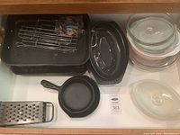 Full lot view showing cast iron fry pans, roasting pans, metal rack, glass casserole dishes, and metal grater.