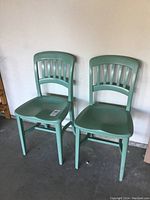 Two vintage painted oak school chairs side by side against a wall on concrete floor.