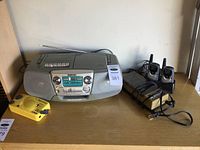 Wide view showing Sony CFD-V7 boombox, Omega Sungear yellow solar radio, Archer battery charger, and Audiovox walkie-talkies with charging base on a wooden surface.