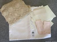 Photo showing folded beige crocheted tablecloth on top of a white fabric tablecloth with stack of six white napkins and two pink cloth napkins beside them.