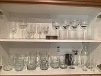 Wide shot on shelf with multiple clear wine glasses, mugs, cocktail glasses and textured bar glasses on two shelves.