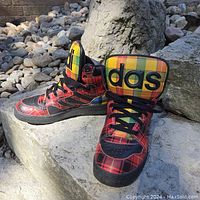 Front and side view of the Adidas Jeremy Scott multi tartan high-top sneakers on rock surfaces, showing colorful plaid patterns with black laces and soles.