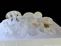 Full view of the assorted crystal and glass serving dishes and decorative items on white tablecloth, showing plates, bowls, candy jar, silver basket, and wire plate stand.