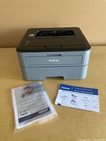 Front and top view of the Brother HL-L2320D laser printer with owner's manual and quick setup guide laid out on table.