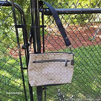 Beige Coach shoulder purse hanging on a chain-link fence showing fabric logo pattern and dark leather strap with exterior zippered pocket.
