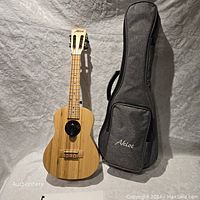Front view of the bamboo tenor ukulele standing upright next to its padded gray carrying case embroidered with 'Aklot'.