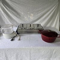 All three items displayed side by side on a white tablecloth: Bravo stainless steel food warmer with three compartments and lids, electric steamer with clear basket, and red Kirkland Dutch oven with lid.