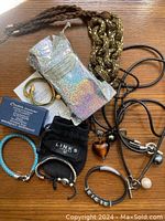 Photo showing assorted bracelets and necklaces including braided leather cords, gold-tone cuff, pendants, and original packaging.