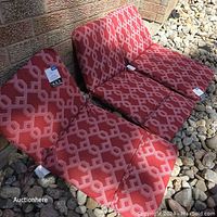 Six red high back patio chair cushions with a geometric pattern laid out on rocks, showing tags attached.