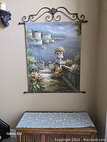 Full view of painted Mediterranean seaside scene wall art hanging on wrought iron decorative hanger on a beige wall above a cabinet.