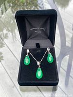 Full set of pendant and earrings in open black jewelry box with brand logo inside lid, showing green teardrop stones and crystal accents.