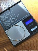 Steel cable bracelet displayed on a digital scale showing a weight of 3.71 grams. The bracelet has a steel twisted wire design with a yellow gold clasp and three gold beads. Visible red enamel detail by the clasp.