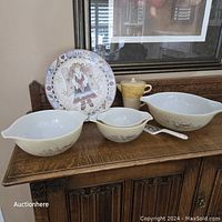 Front view of three vintage Pyrex mixing bowls with beige and brown floral pattern, ceramic cake plate with angel and heart motif, and a yellow/beige teapot and cup, arranged on wooden sideboard.