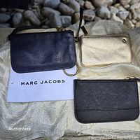 Three assorted zippered pouches displayed on cloth: one black Marc Jacobs with pebbled leather, one metallic gold pouch, and one black Marc Jacobs pouch with white logo text.