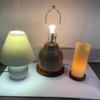 Three lamps on a white surface: a pottery lamp with cream shade, a pottery MCM lamp base on wood, and a frosted glass tube lamp on wood, all lit except MCM without shade.
