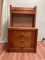 Front view of the teak night stand showing three drawers with wooden pulls and a light behind the top shelf area.