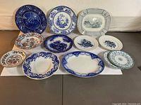 Overview of all 12 vintage decorative plates, bowls, and platters laid out on plain surface showing different colors, sizes, and designs