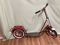 Side view of vintage red dubbed scooter showing full length frame with white wall tires and black grips on handlebars.