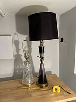 Two lamps on wooden surface, one lamp with black shade and brass accents, the other a clear glass etched base with brass hardware. Tape measure showing size.