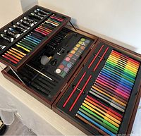 Art box open showing art supplies including acrylic paint tubes, colored pencils, markers, watercolor set, brushes, pencil sharpener