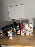 Photo showing 11 assorted spray paint cans in various colors and brands arranged on a wooden surface.