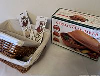 Photo showing two rectangular wicker baskets stacked, with cream fabric liners inside. Also visible are two small decorative metal pieces on cardboard backing, and the boxed German Clay Baker with image of the product on the box.