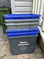 Stacked set of six blue, gray, and one dark gray 'Metro Recycles' outdoor plastic bins with signs of wear and dirt.