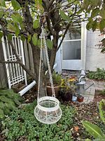 White metal hanging planter suspended outdoors attached to a tree branch, surrounded by garden plants and outdoor items.