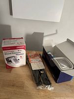 Photo showing Omron Automatic Blood Pressure Monitor box, cuff, and Schweizer magnifier box with magnifiers in plastic packaging on a wooden surface.