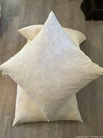 Four light beige or off-white feather pillows stacked, showing clean surface and squared shape.