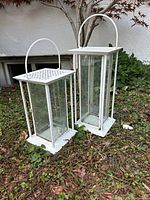 Two white metal and glass lanterns placed outdoors on grass with a small tree in background. Shows overall shape, size, and condition.