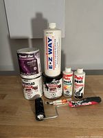 Photo of various paint and cleaner containers along with a paint roller and brush on a gray surface against a gray background.